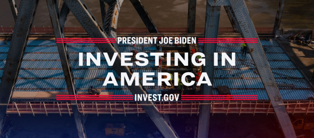 Präsident Joe Biden vor einer Brücke mit der Aufschrift "In America investieren" in der Mitte, umgeben von verschiedenen Gegenständen auf dem Boden.