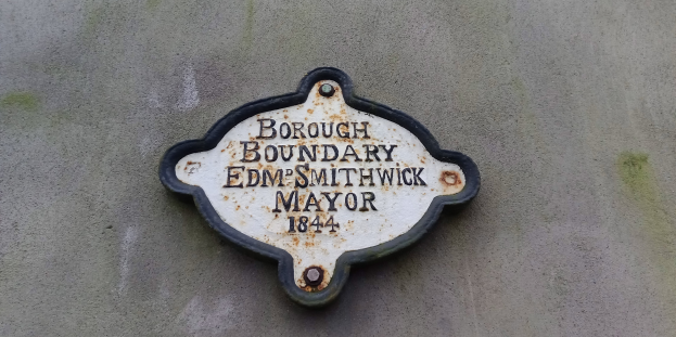 Metallschild an einem Gebäude mit schwarzem Hintergrund und weißer, fetter, zentrierter Schrift, die "Stadtteilgrenze Edm Smithwick Bürgermeister" liest.
