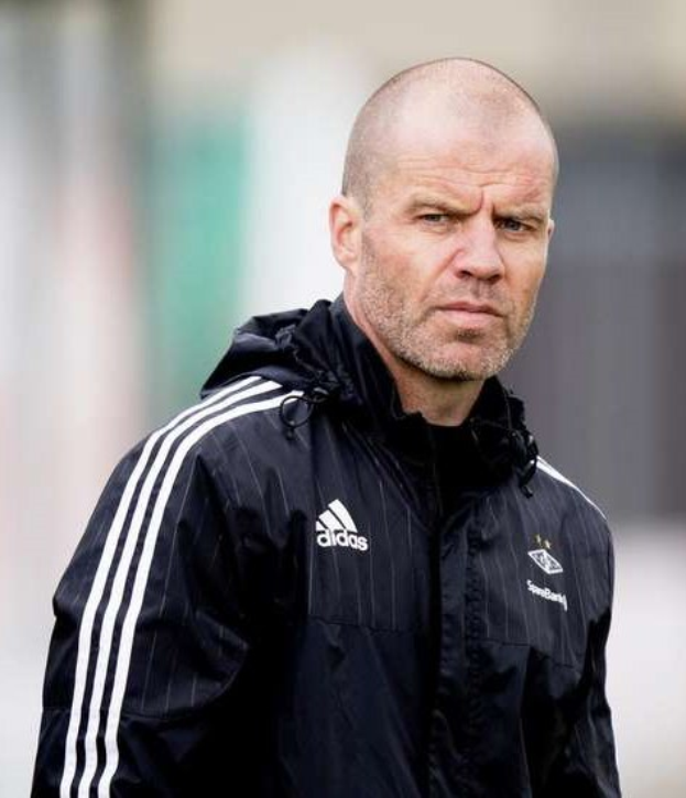 Ein kahlköpfiger Trainer in einer blauen Jacke mit weißen Streifen und weißem Kragen steht auf einem Fußballfeld und schaut ernst in die Ferne.