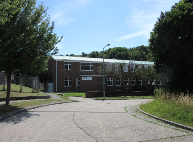 Ein Schulgebäude mit Fenstern, einem Straßenmast, einem Schild, einer Baumgruppe, Gras, einem Zaun und einem bewölkten Himmel, das in der Mitte eines Parks umgeben von saftigem Grün liegt.