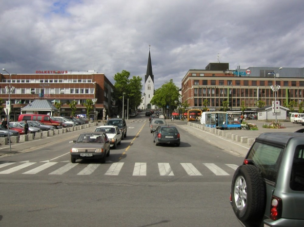 Stadtstraße mit parkenden Autos, Gebäuden, Bäumen, Laternen und einem bewölkten Himmel, mit sichtbarem Kfz-Kennzeichen.