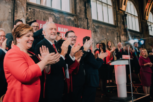 Eine Gruppe von Menschen, die vor einem Publikum applaudieren, mit einem Podium, einem Mikrofon und einer Tafel mit Text rechts daneben, sowie Stühlen, einer Fahne, einer Wand, Fenstern und Lichtern im Hintergrund.