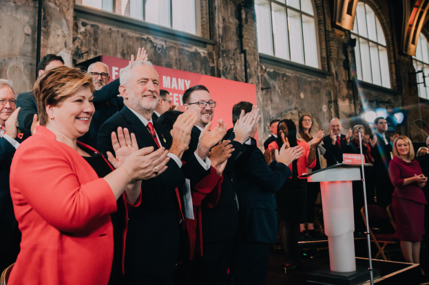 Eine Gruppe von Menschen, die vor einem Publikum applaudieren, mit einem Podium, einem Mikrofon und einer Tafel mit Text rechts daneben, sowie Stühlen, einer Fahne, einer Wand, Fenstern und Lichtern im Hintergrund.