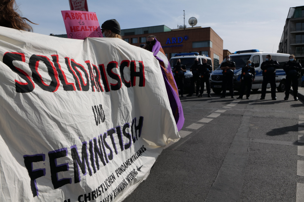 Eine Gruppe von Menschen marschiert auf einer Straße, die ein Banner trägt, auf dem 'Solidarität und Feminismus' steht, mit parkenden Fahrzeugen und Gebäuden im Hintergrund unter einem klaren blauen Himmel.