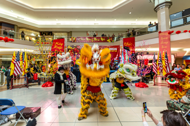 Ein lebendiges chinesisches Neujahrsfest in einem Einkaufszentrum mit Menschen in Kostümen, die tanzen, anderen, die auf Stühlen sitzen und Handys benutzen, mit Fahnen, Bannern, Dekorationen, Geländern, Säulen und Deckenleuchten im Hintergrund.