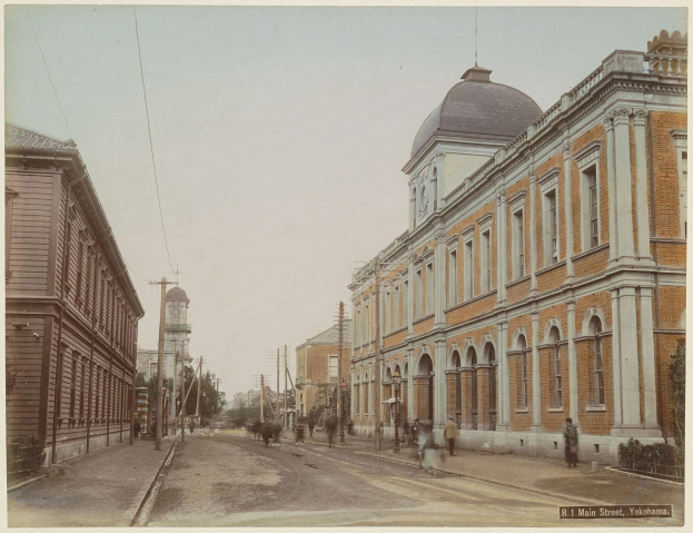 Altes Schwarz-Weiß-Foto einer Stadtstraße mit Gebäuden, Versorgungsmasten, Oberleitungen, parkenden und fahrenden Fahrzeugen, Fußgängern auf dem Gehweg, Bäumen und Himmel mit Text am unteren Rand.