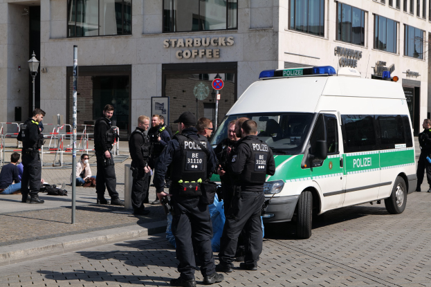 Eine Gruppe von Polizisten steht vor einem Starbucks-Café, rechts ein Van und links ein paar Menschen; im Hintergrund sind ein Gebäude, ein Schild, ein Lichtmast und ein Zaun zu sehen.