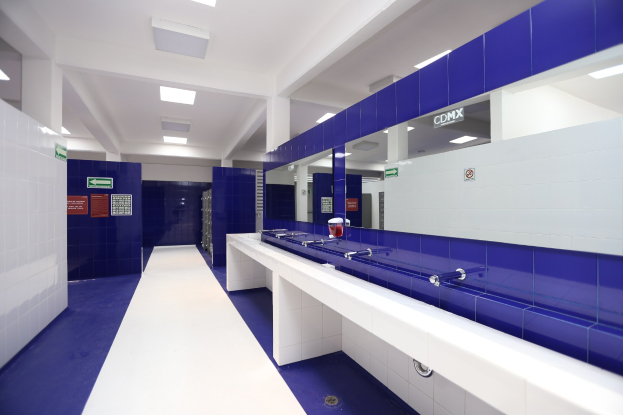 Öffentliche Toilette mit blauen Kacheln, weißen Tresen, Waschbecken mit Wasserhähnen, einer Plattform mit einer Flasche und anderen Gegenständen, Texttafeln an den Wänden, Deckenleuchten und tragenden Säulen.