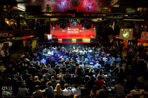 Eine große Menschenmenge sitzt auf Stühlen vor einer Bühne mit Bildschirmen und Bannern, die von Scheinwerfern beleuchtet wird und von Geländern umgeben ist, und blickt erwartungsvoll auf ein Online-Marketing-Rockstars-Event, mit einem Wasserzeichen in der linken unteren Ecke.