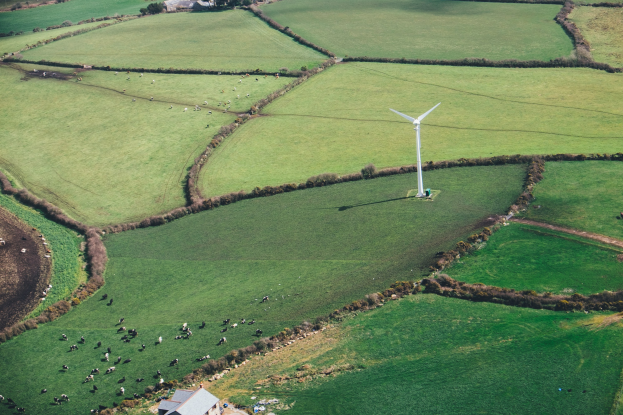 Luftaufnahme eines Windrades auf einer grünen Wiese mit Bäumen, Häusern und Tieren im Hintergrund, das sich in Irland befindet.