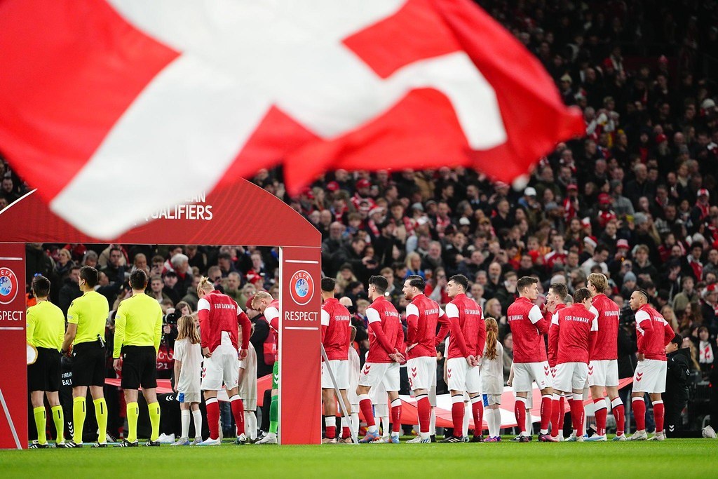 Gruppe von Menschen auf einem Fußballfeld mit einer roten und weißen Flagge im Vordergrund, einem Bogen mit der Aufschrift "Bayern München vs Wetten und Vorschau" im Hintergrund und einer großen Stadionmenge.