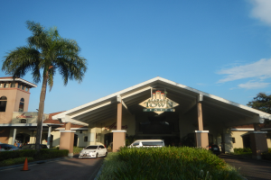 Der Eingang eines Resorts mit Palmen vorne, umgeben von Gebäuden, Kraftfahrzeugen auf der Straße, Verkehrskegel, Büschen, Bäumen und einem bewölkten Himmel im Hintergrund.