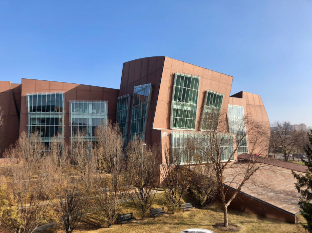 Großes modernes Gebäude mit vielen Fenstern, umgeben von Bäumen, Bänken, Gras und einem Laternenmast, mit anderen Gebäuden und einem klaren blauen Himmel im Hintergrund, beschriftet als der neue Campus der University of Illinois at Chicago.