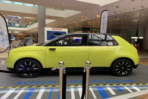 Ein gelbes wasserstoffbetriebenes Elektroauto vor einem Gebäude geparkt, umgeben von Absperrpollern mit Seilen und Schildern, mit einer Person darin, einer Säule, einer Gruppe von Menschen, Schildern, einer Topfpflanze, Deckenleuchten und einer Decke mit Deckenventilatoren.