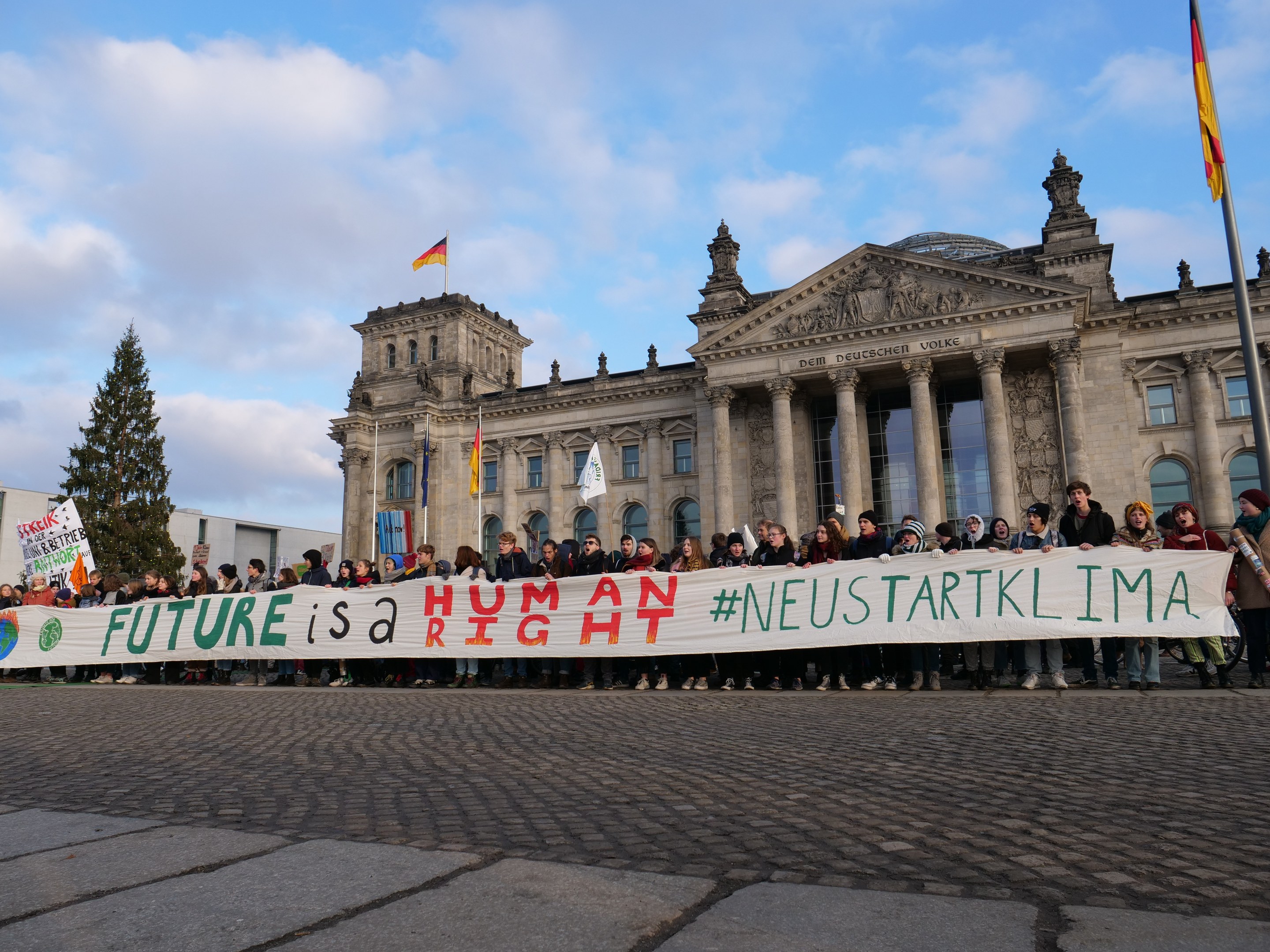 Gruppe von Menschen mit einem Banner 'Zukunft ist ein Menschenrecht' vor dem Reichstaggebäude in Berlin, mit Säulen, Fenstern und Bögen, umgeben von Bäumen und Fahnenmasten unter einem bewölkten Himmel.