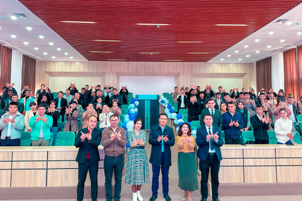 Eine Gruppe von Menschen steht vor einer Menge, einige klatschen, mit Stühlen, Tischen, Ballons, Vorhängen und einer Wand im Hintergrund und Lampen an der Decke, feiernd die Eröffnung einer neuen Schule.