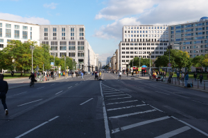 Eine belebte Straßenkreuzung in Berlin, Deutschland, mit Fußgängern und Radfahrern auf der Straße, hohen Gebäuden, Bäumen, Laternenmasten und Schildern unter einem bewölkten Himmel.