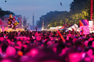 Eine gro├če Menge Menschen auf einem nächtlichen Musikfestival, beleuchtet von bunten Lichtern, mit Zelten, Fahnen, Masten und anderen Gegenständen im Vordergrund und Bäumen, einem Turm und einem klaren Nachthimmel im Hintergrund.