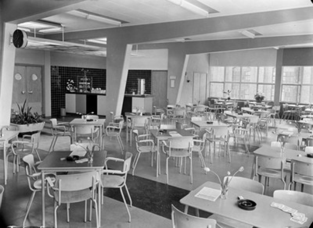 Schwarzes und weißes Foto einer Cafeteria mit Tischen und Stühlen, Tellern, Besteck, Gläsern und Blumenvasen auf den Tischen, Türen und Fenstern an der Wand im Hintergrund und Deckenlampen.