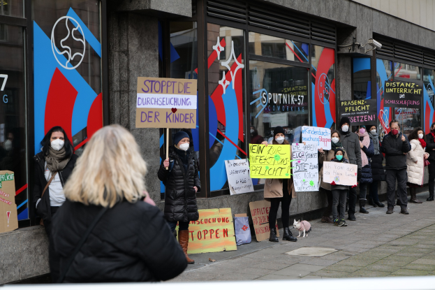 Gruppe von Menschen mit Masken, die Schilder und Plakate vor einem Gebäude mit Glaswänden und -türen halten, während Kameras und ein Hund anwesend sind und gegen das Verbot von Masken in Schulen durch die deutsche Regierung protestieren.
