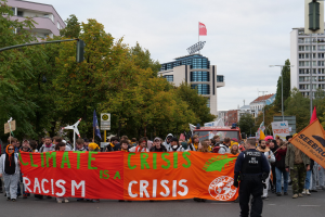 Eine Gruppe von Menschen marschiert eine von Bäumen gesäumte Straße entlang und hält ein Banner in die Höhe, auf dem "Klimakrise ist eine Krise" steht, mit Gebäuden und einem klaren blauen Himmel im Hintergrund.