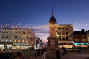 Eine belebte Stadtstraße bei Nacht mit Menschen, Fahrzeugen, Fahrrädern und Gebäuden, beleuchtet von Weihnachtslichtern mit einem Uhrenturm im Hintergrund.