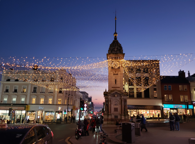 Eine belebte Stadtstraße bei Nacht mit Menschen, Fahrzeugen, Fahrrädern und Gebäuden, beleuchtet von Weihnachtslichtern mit einem Uhrenturm im Hintergrund.