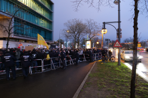 Eine große Gruppe von Menschen vor einem Gebäude mit Schildern, Barrikaden, Fahrrädern, Laternenmasten, Schautafeln, Bäumen und Gras auf dem Boden, unter einem Himmel im Hintergrund, was auf eine Demonstration in Berlin hindeutet.