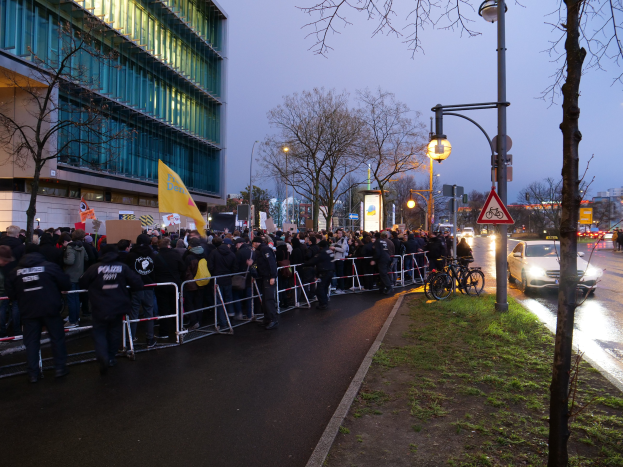 Eine große Gruppe von Menschen vor einem Gebäude mit Schildern, Barrikaden, Fahrrädern, Laternenmasten, Schautafeln, Bäumen und Gras auf dem Boden, unter einem Himmel im Hintergrund, was auf eine Demonstration in Berlin hindeutet.