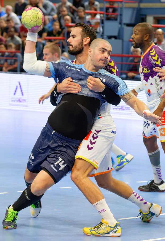 Eine Gruppe von Männern spielt Handball in einem Stadion, wobei ein Spieler den Ball hält.