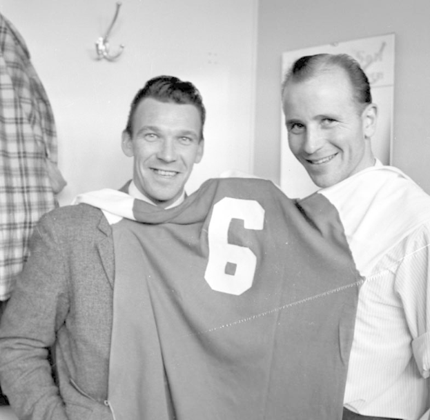 Zwei Männer in Schwarz-Weiß, die strahlend ein Fußballtrikot hochhalten, mit einer Wandtafel und einem Haken im Hintergrund.