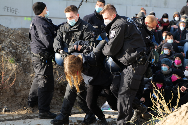 Gruppe maskierter Menschen in Kapuzenpullis, einige mit Waffen, umgeben eine Frau, die von der Polizei eskortiert wird, in einer städtischen Umgebung mit Pflanzen, Steinen und einer Wand mit Protesttext.