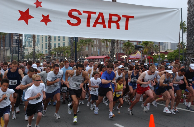 Gruppe von Läufern bei einem Marathon mit einem Verkehrskegel im Vordergrund und einer Fahne im Hintergrund, vor Bäumen, Laternenmasten, Gebäuden und einem klaren blauen Himmel