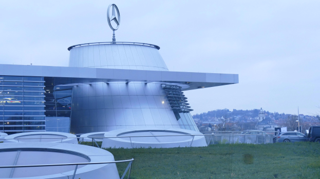 Das Mercedes-Benz Museum in Frankfurt, Deutschland, ein großes Gebäude mit einem Logo darauf, umgeben von Gras und Geländern, mit Fahrzeugen auf der Straße davor und Bäumen unter einem klaren blauen Himmel im Hintergrund.