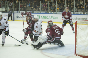 Gruppe von Menschen, die Hockey auf einem Eisstadion spielen, mit Torpfosten auf der rechten Seite, tragen Helme und halten Hockeystöcke, Zuschauer auf den Tribünen mit Bannern im Hintergrund.