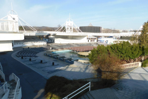 Blick auf das Olympiastadion von einem hohen Standpunkt aus, mit verschiedenen Objekten und Grün im Vordergrund und einem bewölkten Himmel im Hintergrund.