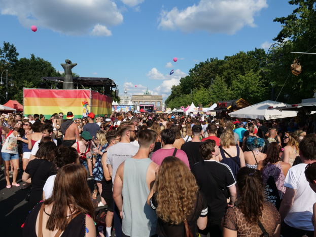 Eine große Menschenmenge geht eine Straße mit Zelten, Bäumen, Pfählen, Lichtern und einer Statue entlang, mit Gebäuden und einem Himmel voller Wolken und Ballons im Hintergrund während des Christopher Street Day in Berlin.