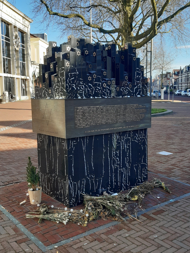 Ein Holocaust-Denkmal in einem Stadtplatz, mit einem Baum davor, einer Topfpflanze auf dem Boden und verstreuten trockenen Blättern, sowie Gebäuden, Fahrzeugen, Pfählen und Bäumen im Hintergrund.