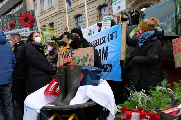 Maskierte Menschen mit Protestschildern vor einem Lastwagen mit einem Tisch, Pflanzen und einer Flagge im Hintergrund.