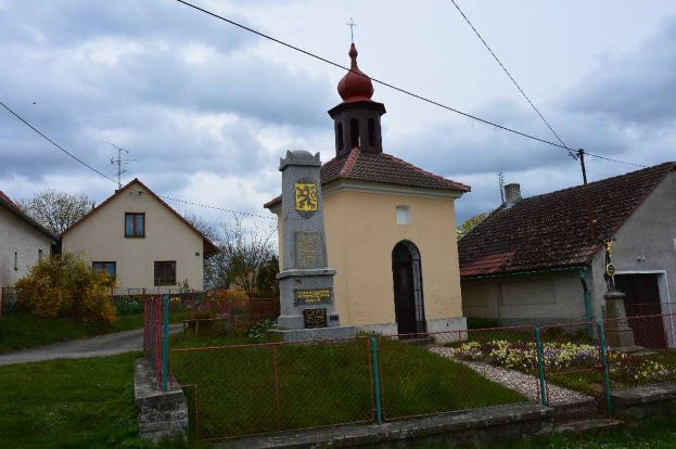 Kleine Kirche mit einem Glockenturm als Kirche des Heiligen Grabes umgeben von Häusern, Grünflächen, einem Zaun und einem bewölkten Himmel.