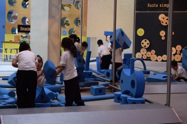Kinder spielen in einer Turnhalle und halten Gegenstände, mit einer Texttafel an der Wand und Stangen rechts.