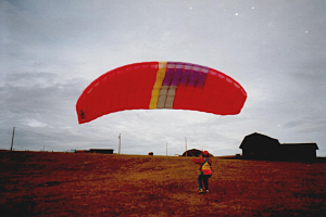 Eine Person mit einem roten Fallschirm Gleitschirmfliegen in einem Feld, umgeben von Häusern, Pfählen und einem klaren blauen Himmel.