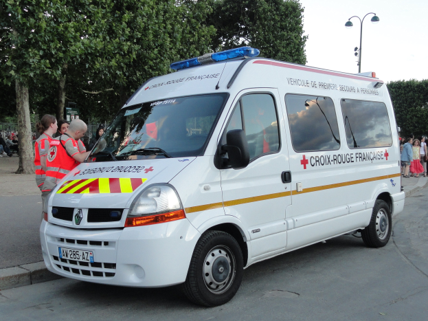 Ambulanz am Straßenrand mit einer Gruppe von Menschen in roten Jacken in der Nähe, Bäume und Laternen im Hintergrund unter einem klaren blauen Himmel.