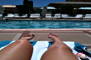 Frauen auf Liegestühlen am Poolrand mit Handtüher, mit Blick auf ein Schwimmbad, umgeben von Bühschen und Häusern.