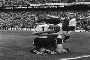 Drei Fußballspieler umarmen sich auf dem Boden, feiern ein Tor vor einer jubelnden Menge in einer schwarz-weißen Stadionszene.