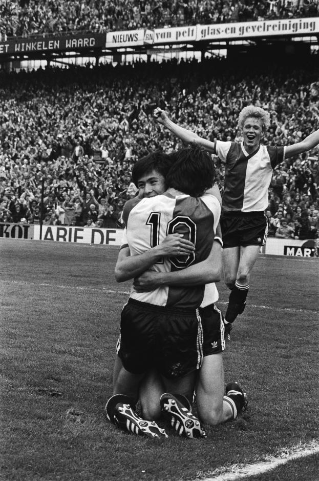 Drei Fußballspieler umarmen sich auf dem Boden, feiern ein Tor vor einer jubelnden Menge in einer schwarz-weißen Stadionszene.