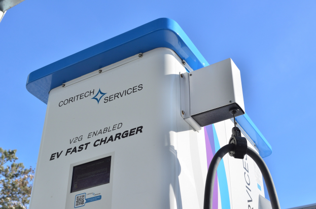 Elektrofahrzeug-Ladestation mit einem blauen und weißen "V2G Fast Charger"-Schild, umgeben von Bäumen und einem Gebäude unter einem klaren blauen Himmel.