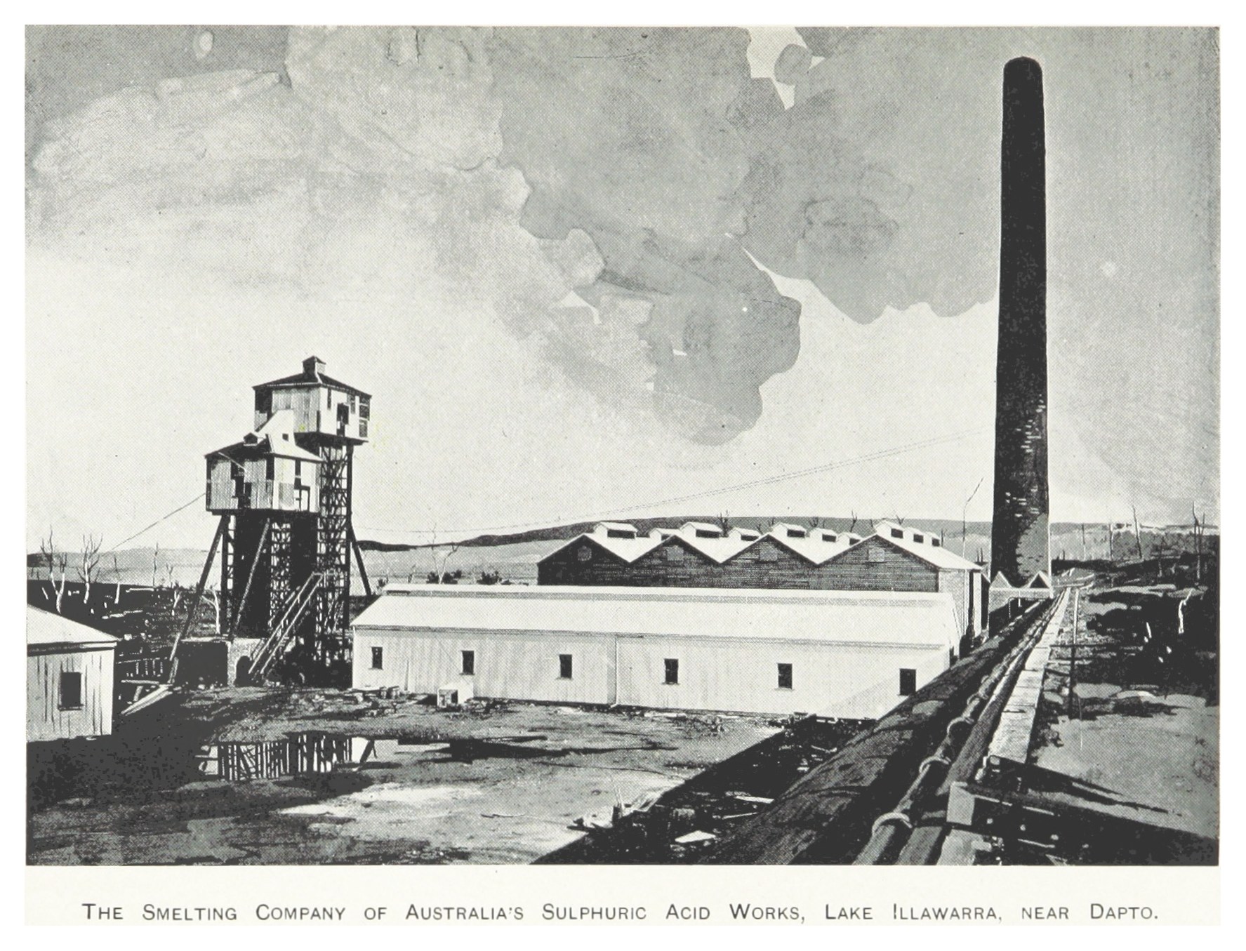 Schwarzes und weißes Foto einer Industrieanlage mit mehreren Gebäuden, einem hohen Turm und Rauch, der aus einem Schornstein aufsteigt, mit Text unten.