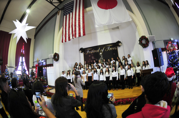 Gruppe von Menschen auf einer Bühne mit Weihnachtsdekorationen, einem Rednerpult, Lautsprechern, einer Texttafel, Vorhängen, Fahnen und Deckenleuchten vor einem Publikum mit Kameras und Handys.