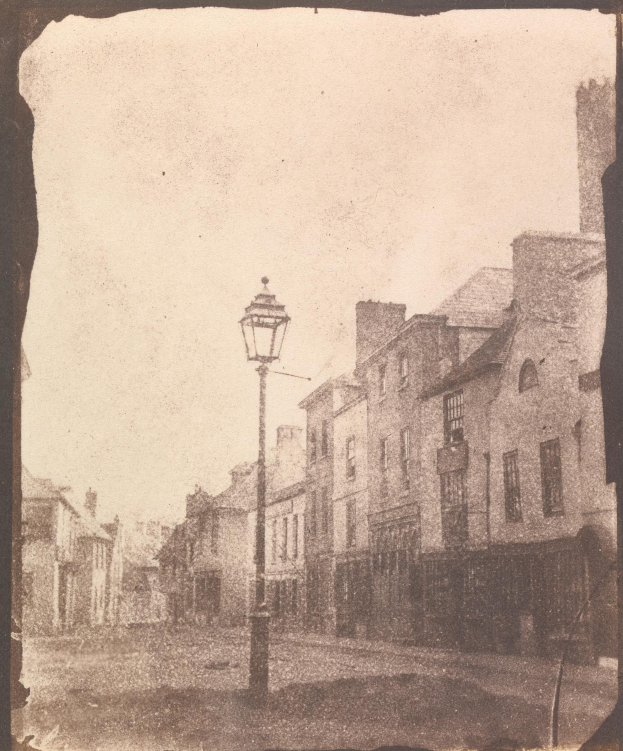 Altes Schwarz-Weiß-Foto einer Straßenlaterne an einer Kreuzung, flankiert von Gebäuden unter einem klaren Himmel.